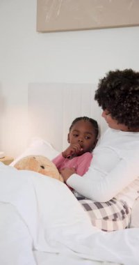 Afro-Amerikan anne ve kızı yatakta saç okşuyor hap yerleştiriyor, telefonla konuşuyor. Bakım, bakım, rahatlık, şefkat, uyku zamanı, iyileştirme, dikey video
