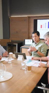 Doğu Asyalı adam el kol hareketi yapıyor, sunucu ekrandaki grafiğe işaret ediyor ve veri eğilimlerini açıklıyor. İşbirliği, takım çalışması, analitik, iş toplantısı, profesyonel, modernlik, dikey video