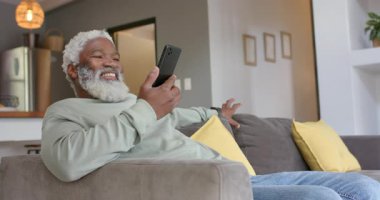 Olgun bir Afro-Amerikalı adam kolçaktan akıllı telefonu kaldırıyor ve gri kanepede haber için kayıyor. Teknoloji, rahatlama, oturma odası, gündelik, modern, konfor, yaşam tarzı