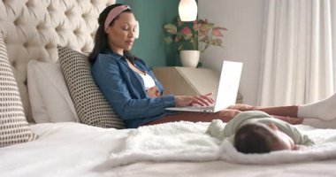 Anne yatak odasındaki battaniyedeki bebeği izlerken dizüstü bilgisayarı kucağına yerleştiriyor ve yatakta daktilo yazıyor. Bakıcı, aile, rahatlama, evcil, rahat, besleyici, modern