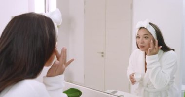 Cilt bakımına başlayan sıradan bir Asyalı kadın nemlendirici kavanozuna uzanıyor, nemlendirici sürüyor. Cilt bakımı, güzellik, kendine bakım, hijyen, dinginlik, minimalizm, sağlık