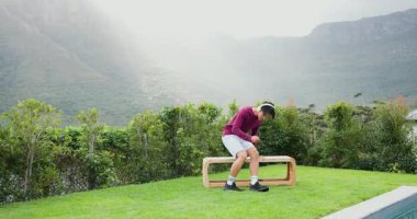 Bahçeye giren Asyalı adam akıllı telefon kullanarak bankta oturuyor tepeleri işaret eden dağları seyrediyor. Açık hava, sükunet, modern, yaşam tarzı, doğa, rahatlık, manzara