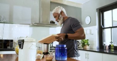 Afrika kökenli Amerikalı kıdemli bir adamın mutfakta meyve püresi için muz ve protein tozu karıştırması. Sağlıklı, beslenme, yemek pişirme, yaşam tarzı, modern, fitness, sağlık