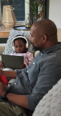 Dikey video: Afro-Amerikalı baba koltukta pelüş oyuncağı ve telefonu tutuyor ve kızını güldürüyor. Aile, bağ, teknoloji, rahatlama, neşe, ev, dikey video