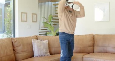 Olgun Asyalı bir adam evdeki koltukta beyaz sanal gerçeklik kulaklığını fark ediyor ve oynamadan önce takıyor. Teknoloji, daldırma, yenilik, eğlence, eğlence, minimalist, dijital