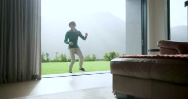 Kulaklıklı Asyalı adam odaya giriyor, dans ediyor, tablet akıllı telefonu karşılaştırıyor, kanepede dinleniyor. Kişisel teknoloji, eğlence, modern tasarım, yaşam tarzı, rahatlama, aktivite, kapalı alan