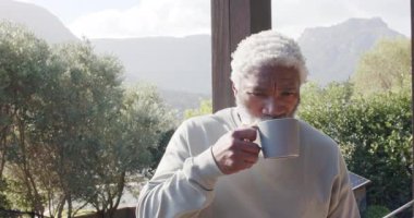 Kıdemli Afro-Amerikan adam balkona çıkıyor, bardak kaldırıyor, yudumluyor ve dağ manzarasına hayran kalıyor. Dalgınlık, huzur, doğa, sağIık, farkındalık, kırsallık, manzara