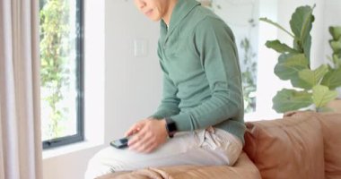 Asyalı adam oturma odasındaki koltukta akıllı telefon haberlerini bekliyor, sonra telefona sarılıp, tezahürat yapıyor. Modern, gündelik, neşeli, teknoloji, iç mekan, rahatlama, ifade