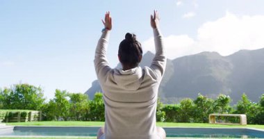 İçeriye odaklanmak için kollarını havaya kaldıran Asyalı kadın havuz başı yoga minderinin üzerinde dağlara bakıyor. Meditasyon, dinginlik, farkındalık, sağlık, açık hava, manzara, rahatlama.