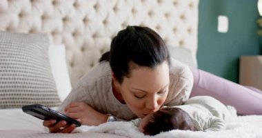 Anne ve bebek yatakta, anne akıllı telefon tutuyor. Bebeği öperken hafızasını yakalıyor. Aile, bakım, yakınlık, rahatlık, bağlantı, doğal ışık, sıcak ortam