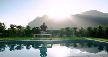 Asyalı adam, havuzun yanındaki tahta bankta meditasyon yapmadan önce kollarını kaldırıp güneşin doğuşunu selamlıyor. Dinginlik, farkındalık, ahenk, doğa, sükunet, refah, gündoğumu