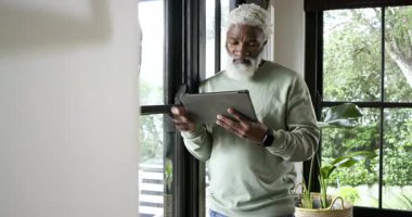 Olgun Afro-Amerikan bir adam pencerenin önünde duruyor ve saksının yanında tablet tarama haberleri sunuyor. Teknoloji, rahatlama, modern yaşam tarzı, açık hava, dinginlik, farkındalık