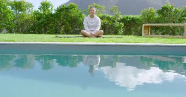 Havuz başına gelen Asyalı kadın yoga minderine oturmuş meditasyon yapıyor ve kollarını başımın üstüne uzatıyor. Zen, sükunet, farkındalık, rahatlama, açık hava, doğa, sağlık