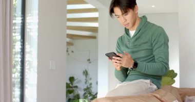 Asyalı adam evdeki koltukta telefonu kaydırıyor, içeriğe tepki veriyor, geri düşüyor, yükseliyor, yumruk sallıyor. Gündelik, rahatlama, yaşam tarzı, modern, anlamlı, canlı, rahat