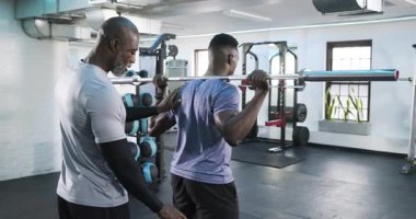 Afrika kökenli Amerikalı erkek eğitmen, stajyer halter askılığına rehberlik ediyor, inişini ve çıkışını güvenli bir şekilde görüyor. Fitness, koçluk, güç, istikrar, eğitim, motivasyon, dayanıklılık