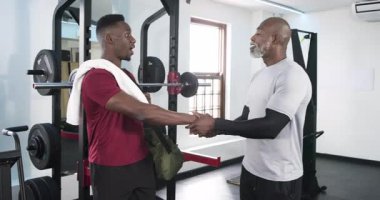 Afrikalı Amerikalı antrenör ve müşteri spor salonuna spor çantasıyla giriyor, tasmaları ayarlamadan önce el sıkışıyorlar. Zindelik, güç, egzersiz, eğitim, motivasyon, sağlık, sağlık