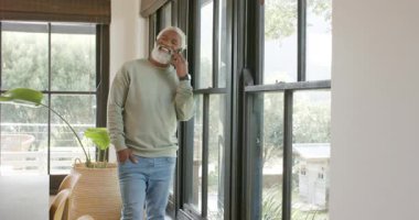 Afro-Amerikalı bir adam oturma odasına giriyor akıllı telefonu camdan kulağa kaldırıyor, ararken gülümsüyor. Kentsel, güncel, çağdaş, yaşam tarzı, rahatlama, iletişim, ev