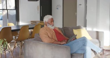 Kanepeye girip yerleşen olgun Afro-Amerikan bir adam oturma odasındaki tablet haberlere kayıyor. Yaşam tarzı, rahatlama, teknoloji, ev, rahat, rahat, boş zaman