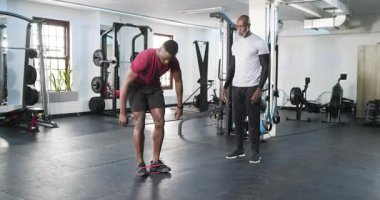 Afrikalı Amerikalı erkek müşteri spor salonunda kırmızı bant takıyor, antrenör gözlemlerken diz yükseltiyor. Spor, güç, rehabilitasyon, kas, dinamik, duruş