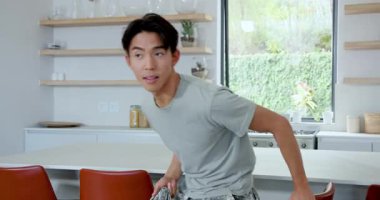 Mutfağa elinde kamuflaj ceketiyle giriyor, Asyalı bir erkek kırmızı taburede oturuyor, onu düzeltiyor, gülümsüyor. Modern, gündelik, yaşam tarzı, iç mimari, modaya uygun, rahat, hayat dolu.