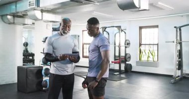 Afrikalı Amerikalı erkek antrenör ve müşteri tablet tıkırdatıyor, spor yapmayı planlıyor ve spor salonunda yumruk tokuşturuyor. Koçluk, fitness, teknoloji, motivasyon, güç, eğitim, takım çalışması