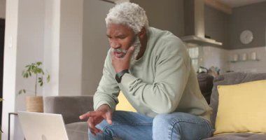 Üst düzey Afro-Amerikan adam dizüstü bilgisayara kaşlarını çatıyor, daktilo ediyor, oturma odasında yardım istiyor. Modern, profesyonel, minimalist, teknoloji, iletişim, gündelik, iç mekan