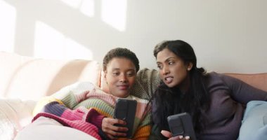 Evdeki kanepede uzanan çeşitli bayan arkadaşlar telefon ekranına gülerek akıllı telefonları kaydırıyorlar. Arkadaşlık, rahatlama, ev ortamı, konfor, bağlantı, çeşitlilik, rahatlık