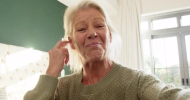 Olgun bir kadın, zeytinli kazağıyla yatak odasında konuşurken elini kalbe koyuyor. Özgünlük, samimiyet, iletişim, farkındalık, sağlık, dinginlik, içgözlem