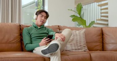 Akıllı telefon uyarısı aldıktan sonra Asyalı bir adam, kulaklıklı bir şekilde kanepeye telefon takıp kayıyor. Gündelik, modern, rahatlama, teknoloji, iletişim, konfor, yaşam tarzı