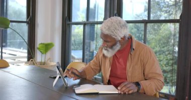 Kıdemli Afro-Amerikalı adam tablet kaydırıyor ve not defterine pencere kenarına yazıyor, kopyalama alanı. Profesyonel, teknoloji, modern, çalışma alanı, okur yazarlık, yaşam tarzı, kentsel