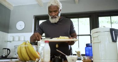 Kıdemli Afro-Amerikalı adam mutfakta muz dilimliyor smoothie için blender 'a karıştırıyor. Mutfak, beslenme, sağlıklı, yaşam tarzı, modern, kırsal, tazelik