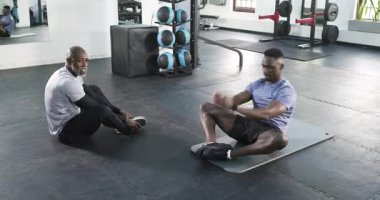 Olgun Afro-Amerikan erkek fitness koçu spor salonunda kalçasını hareket ettirebilmek için paspasa uzanan stajyeri işaret ediyor. Eğitim, esneklik, sağIık, kaporta, güç, teknik, farkındalık
