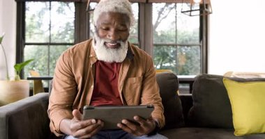 Olgun bir Afro-Amerikan erkeği tablet içeriğine tepki veriyor, kanepeye vurup gülüyor. Teknoloji, rahatlama, rahatlama, modern, yaşam tarzı, konfor, eğlence