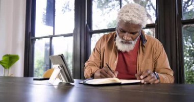 Üst düzey Afro-Amerikan adam planlama için ofis masasında notlar yazarken tablete göz atıyor. Profesyonel, çalışma alanı, minimalist, üretkenlik, yenilikçi, düşünceli, doğadan esinlenilmiş