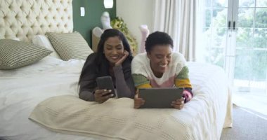 Çeşitli bayan arkadaşlar tablet içeriğine kıkırdıyor ve yatakta yatarken telefon ekranlarını değiştiriyorlar. Arkadaşlık, rahatlama, dijital bağlantı, rahat, gündelik, çeşitlilik, modern