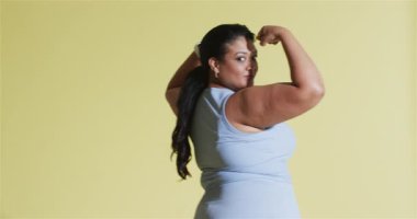 Yetişkin bir kadın, sarı stüdyoda dönüyor. Aktif giyimli, beyaz, akıllı halka küpeler takıyor. Atletik, güç, güven, yetkilendirme, motivasyon, fitness, dinamik