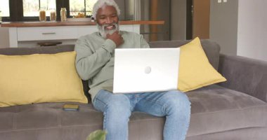 Kıdemli Afrikalı Amerikalı adam oturma odasındaki kanepeye yerleşiyor, gülüyor, çalışmak için dizüstü bilgisayarda yazıyor. Nesilsel rahatlama, teknoloji, konforlu, çağdaş, boş zaman, ev bakımı