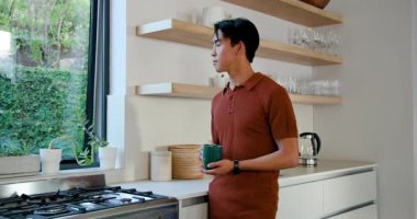 Yetişkin Asyalı adam mutfak tezgahında yeşil bardaktan yudumlarken dışarıya bakıyor ve kokuyu içine çekiyor. Rahat, huzur, minimalist, rahatlama, evcil, yaşam tarzı, sakinlik