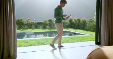 Asyalı adam güneşli terasa çıkıp parlak sabah ışığından kaynaklanan tablet okuyor. Lüks, eğlence, sofistike, modern, açık hava, tefekkür, refah