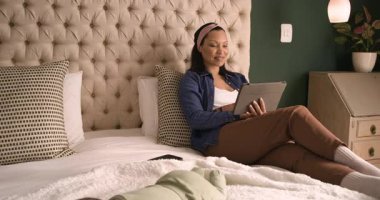 Yatakta tableti tıkırdatan yetişkin bir kadın battaniyedeki uyuyan kızına bakıyor. Annelik, samimiyet, rahatlama, konfor, teknoloji, beslenme, şefkat