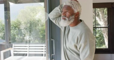 Olgun Afro-Amerikan bir adam cam kapıyı sürgüleyerek arka bahçeye bakıyor ve beyaz bankta fotokopi çekiyor. Düşünce, rahatlama, sükunet, açık hava, ev içi, doğa, yansıtıcı