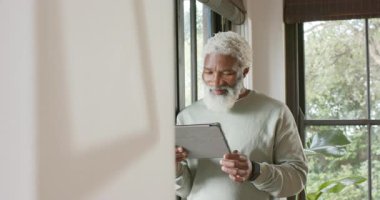 Kıdemli Afro-Amerikan adam, akıllı ayaklı tablet giyiyor, içeriği okuyor ve pencereye bakıyor. Teknoloji, son sınıf, sağlık, yaşam tarzı, rahatlama, dijital, ev