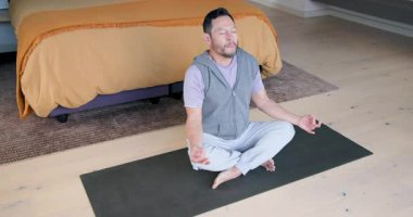 Olgun Asyalı adam yatak odasında yoga minderinde oturuyor, gözlerini kapatıyor, içeriye doğru sükunet işareti veriyor, mudra oluşturuyor. Meditasyon, farkındalık, dinginlik, sükunet, yoga, rahatlama, sağIık