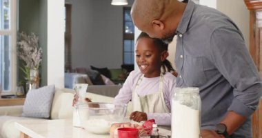 Babası mutfak adasında rehberlik ederken ve gülümserken Afrikalı Amerikalı kız sütü kaseye dolduruyor. Aile, bağlanma, eğitim, yetiştirme, yerli, ev yapımı, pastel