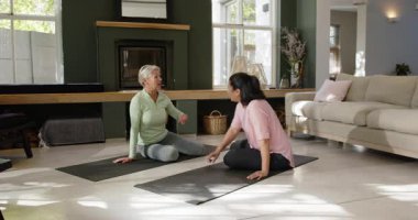 Çeşitli kadınlar oturma odasının zemininde el kol hareketleri yapıp yoga minderlerini yuvarlıyor, yer açıyor. Sağlık, farkındalık, işbirliği, rahatlama, ev, çağdaş, uyum