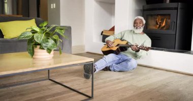 Kıdemli Afro-Amerikalı adam evde şöminenin yanında durup akustik gitar tıngırdatıyor. Rahatlama, müzik, konfor, oturma odası, sükunet, hobi, konfor