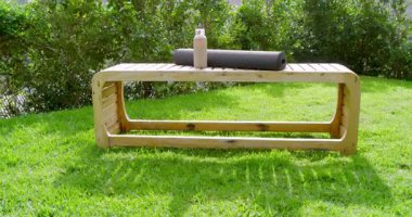 Kamera, yoga minderi ve su şişesiyle çimenlikteki bankı geri çekiyor. Dağları gösteriyor. Manzara, doğa, refah, açık hava, dinginlik, sükunet, yeşillik