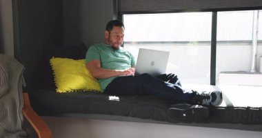 Bilgisayarı pencereyle açtıktan sonra adam daktilo ediyor, dinlenmek için kapanıyor, güneş ışığı odasını doldururken geriniyor. Çalışma alanı, modern, rahatlama, güneş ışığı, iç mekan, gündelik, dijital