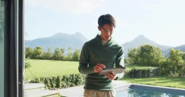 Asyalı bir adam verandaya cam kapıdan giriyor, havuz kenarında yürüyor ve tableti aşırıyor. Açık hava, teknoloji, yaşam tarzı, modern, boş zaman, huzur, manzara