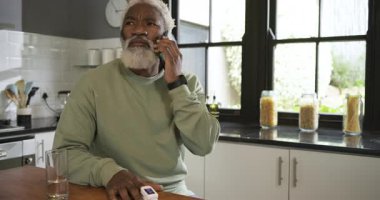 Afro-Amerikalı olgun bir adam mutfakta telefona cevap veriyor ve oksijeni parmak ucu oksimetresiyle test ediyor. Teknoloji, sağlık, sağlık, ev ortamı, modern, yaşam tarzı, sağlık izleme